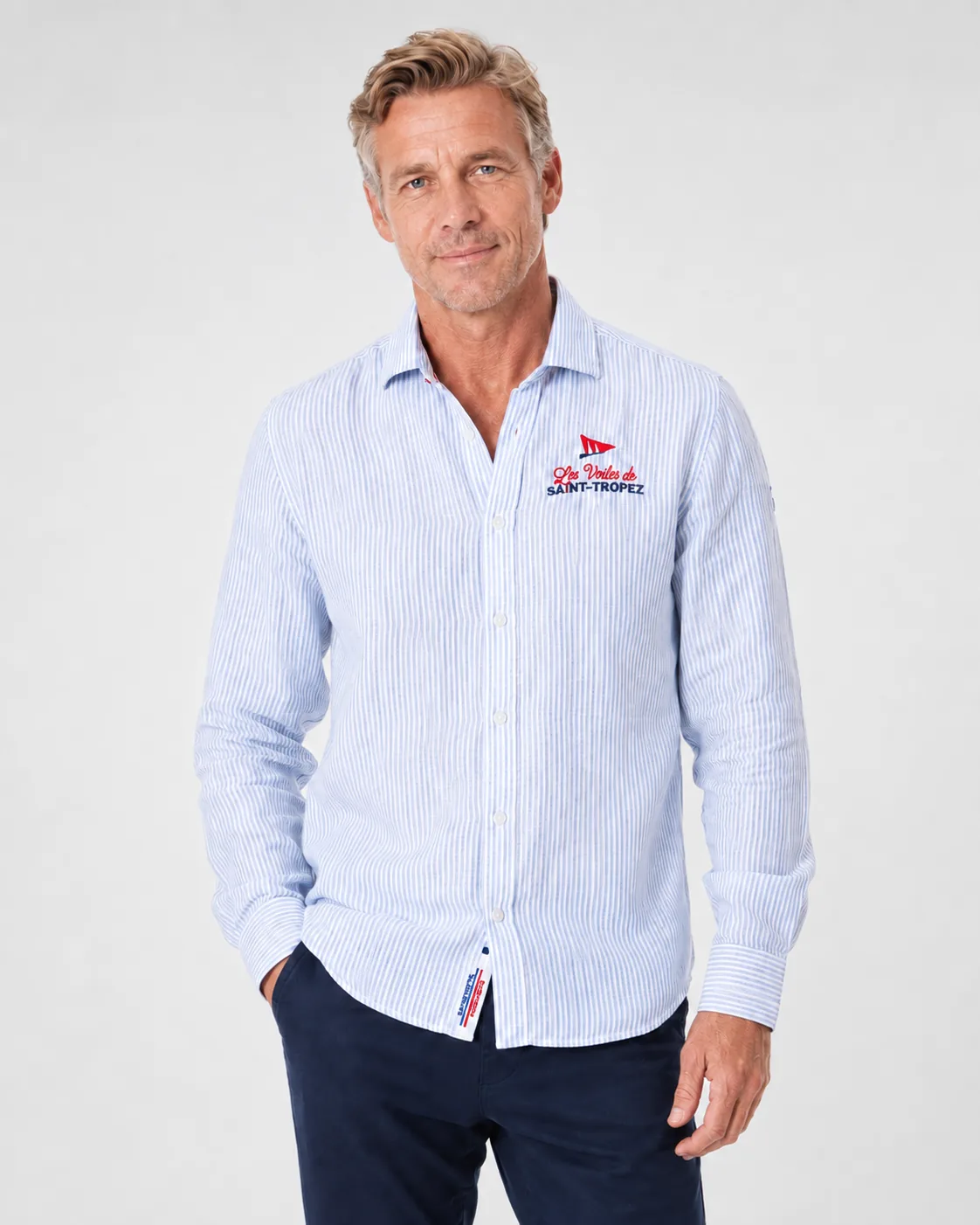 Chemise Lin 26 in a fine sky blue stripe worn from the front: open spread collar and the Les Voiles de Saint-Tropez pennant embroidered on the chest.