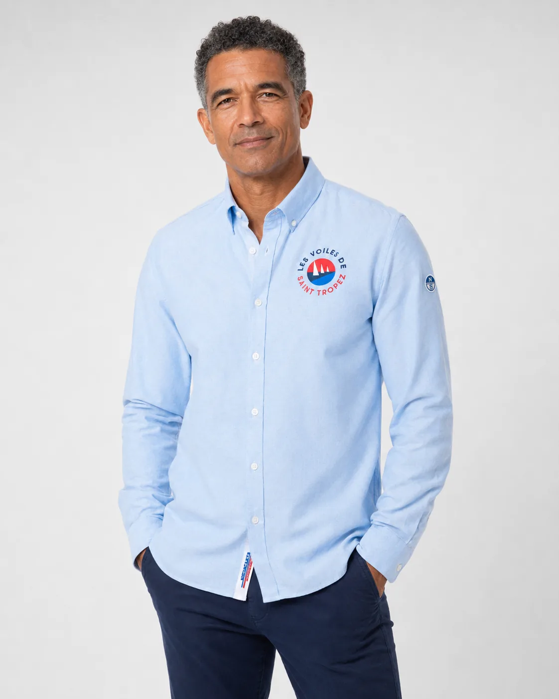 Chemise Oxford 26 in sky blue worn from the front: button-down collar and round Les Voiles de Saint-Tropez badge on the chest.