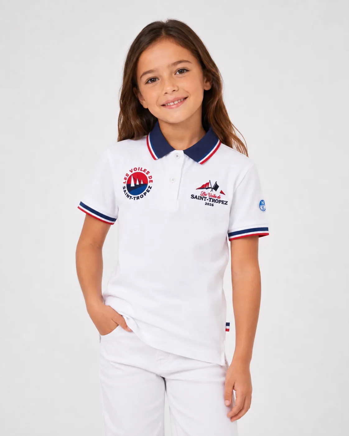 Polo Kids 26 in white worn from the front by a child: navy collar with tricolour tipping, round badge on the left, Les Voiles de Saint-Tropez 2026 lettering on the right and a North Sails patch on the sleeve.