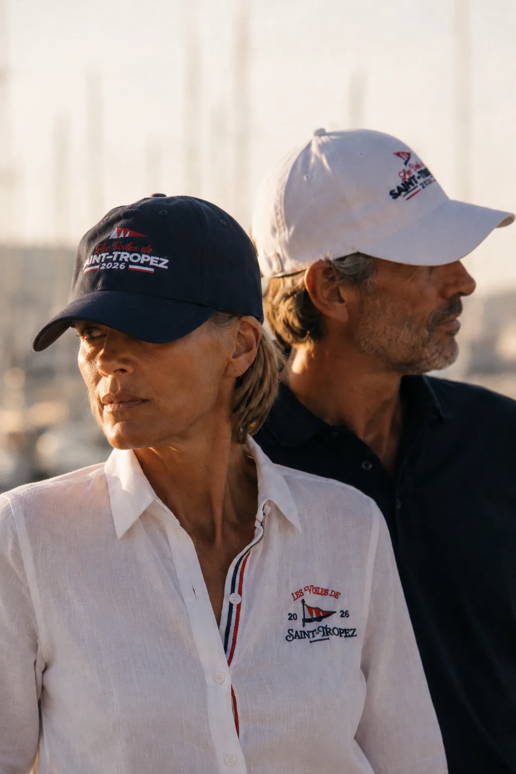 Two people seen from behind at the harbour, wearing navy and white caps embroidered Les Voiles de Saint-Tropez 2026.