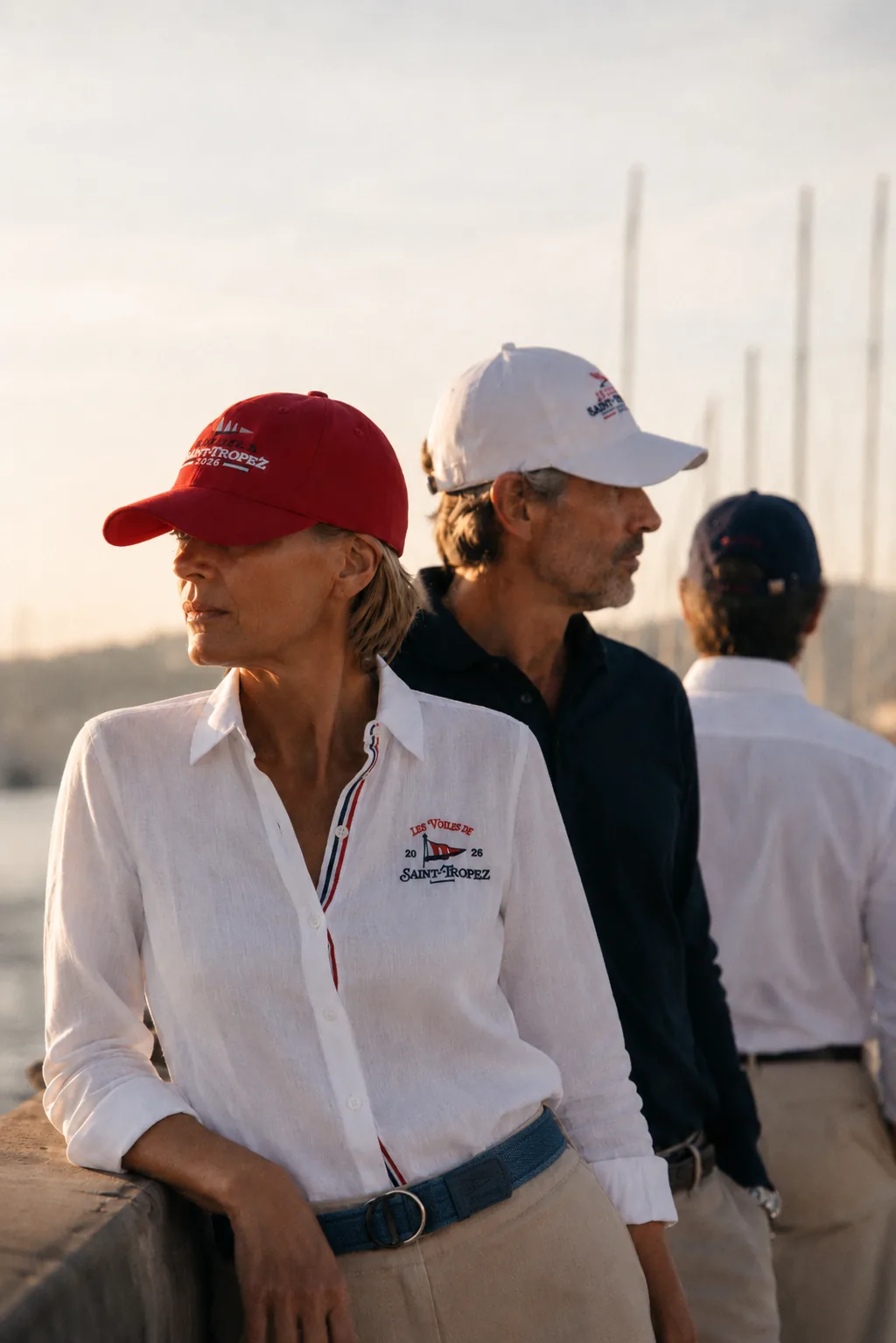 Three people on the quay at sunset; in the foreground, the red cap from the 2026 collection, with a white one and a navy one behind.