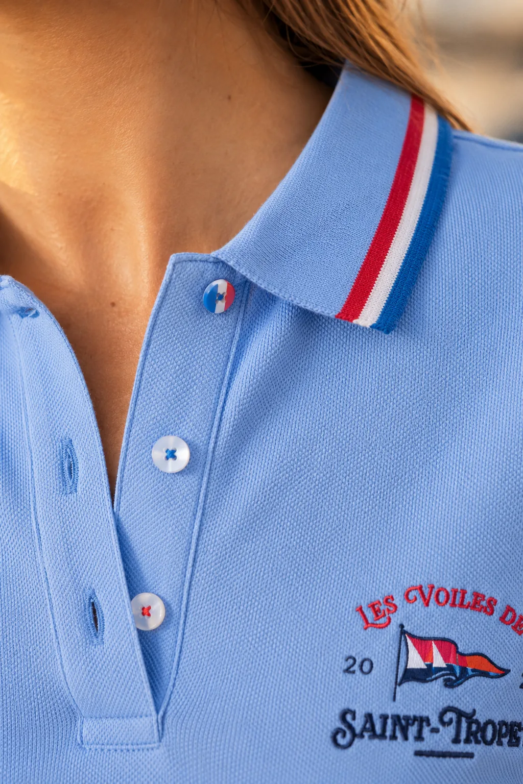 Close-up of the collar and placket of a light blue women's polo shirt: tricolour tape and mother-of-pearl buttons, one of them blue-white-red.