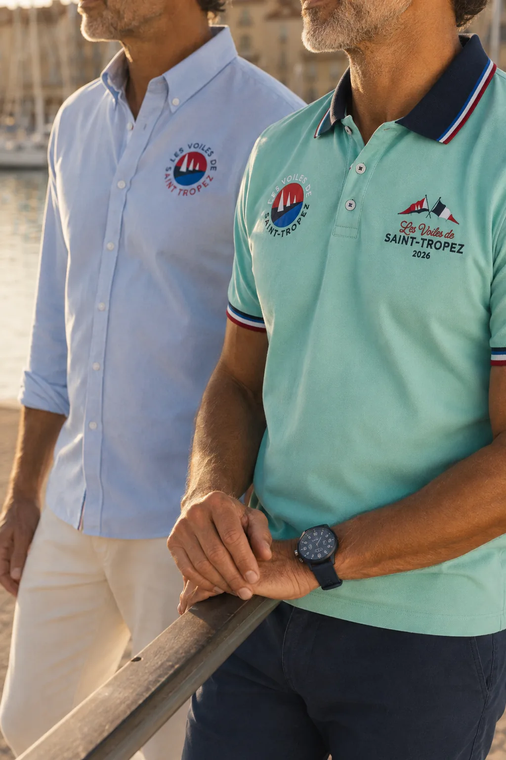 Close-up of two men at the harbour: a light blue Oxford shirt and a mint green polo shirt, with the “Les Voiles de Saint-Tropez 2026” badge embroidered on the chest.