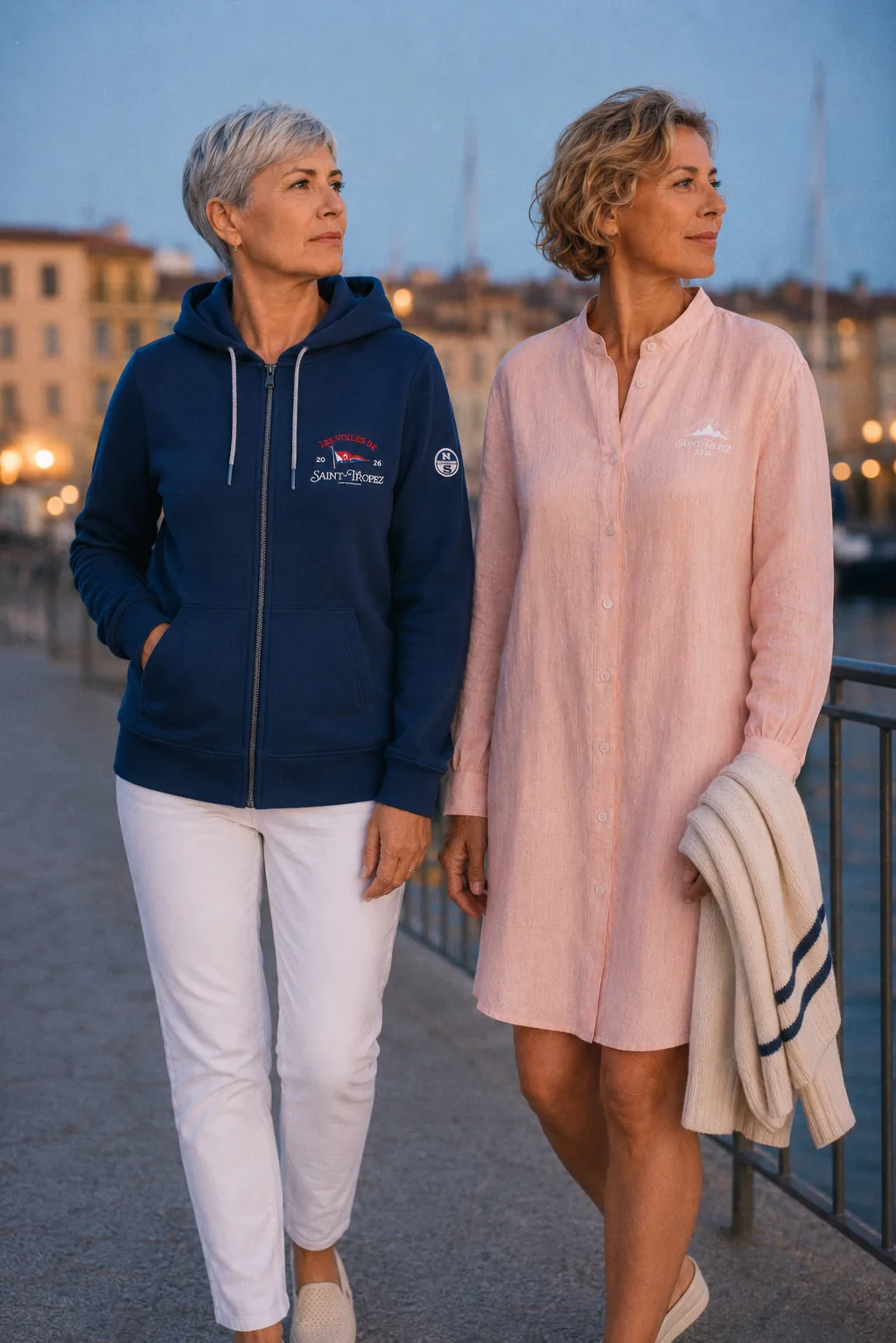 Two women walking along the quay at dusk, in a navy zip-up sweatshirt and a coral pink linen shirt dress.