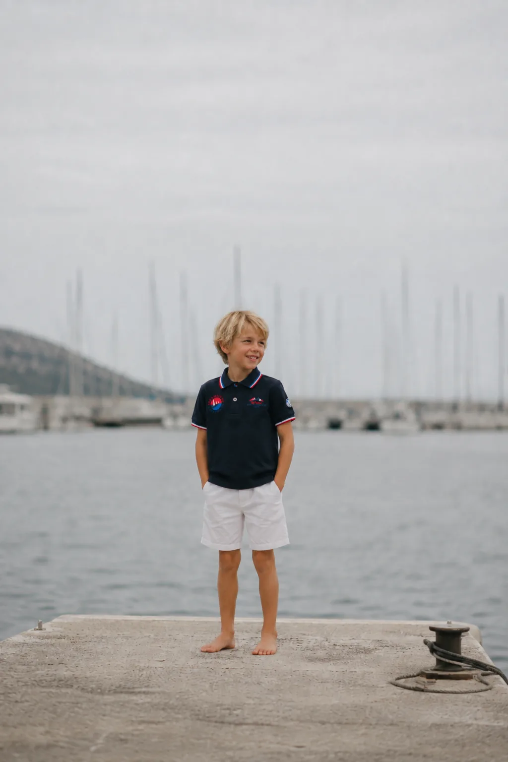 A child in a navy polo shirt from the 2026 collection, standing barefoot at the end of a harbour pontoon.