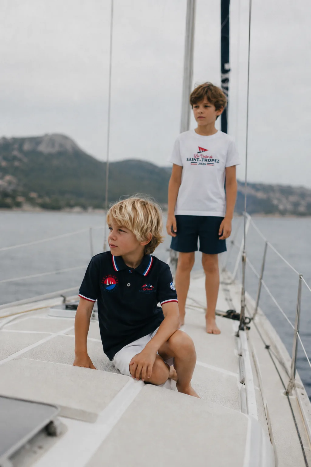 Two children aboard a yacht, in a navy polo shirt and a white T-shirt from the 2026 collection.